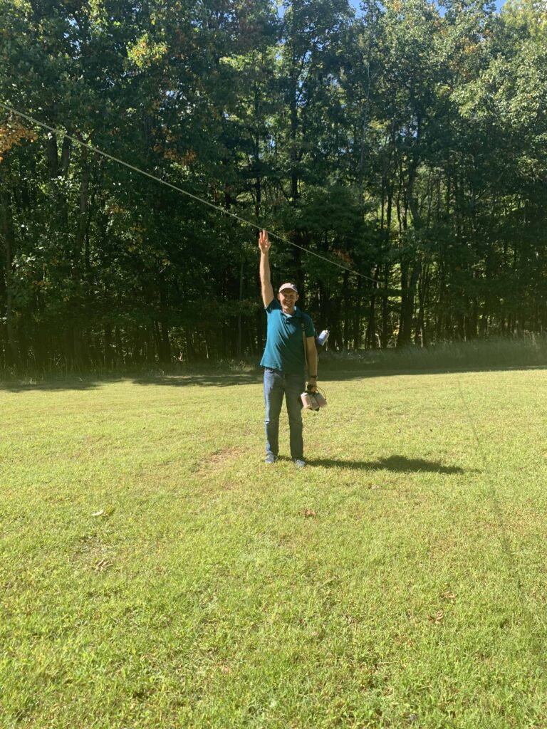 guy standing on grass with his arm up to show how high the zipline is